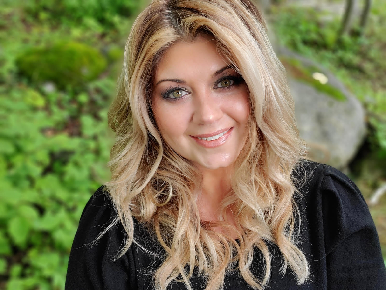 Woman with long blonde hair smiling outdoors with greenery in the background